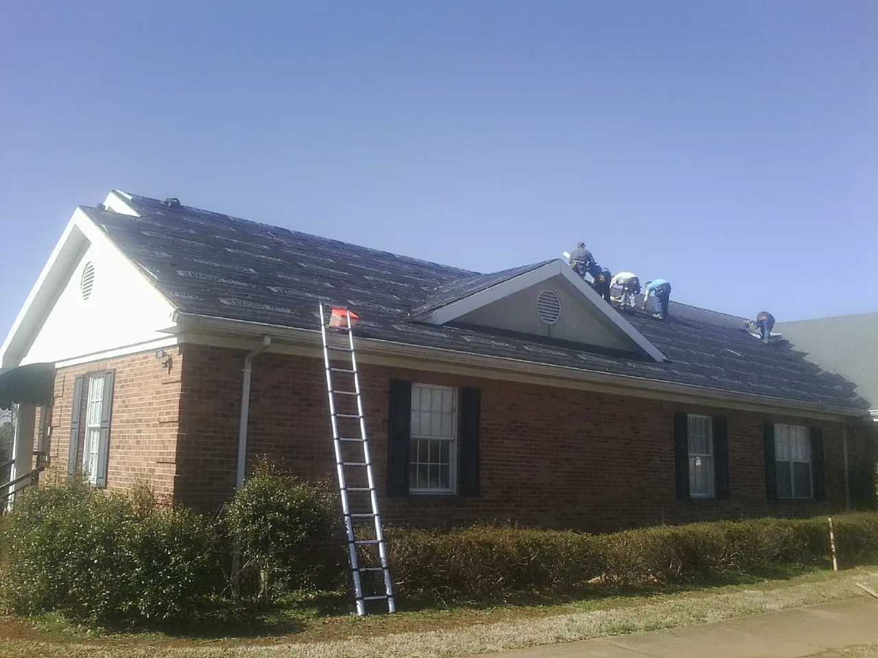 Crew tearing shingles off a brick ranch roof with a ladder set — Metal Roof Repair work by our crew in Hankins