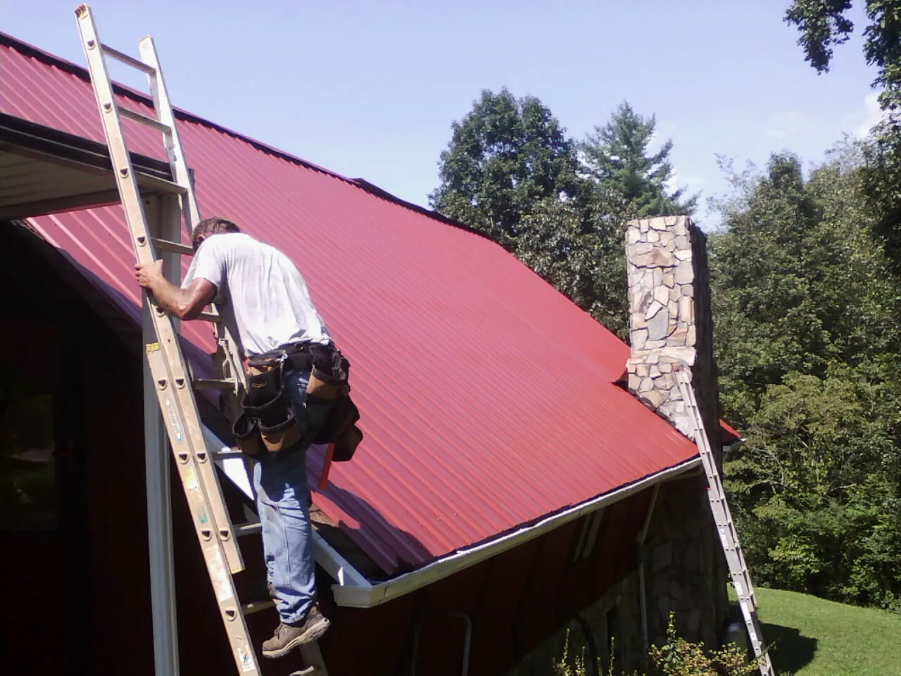 Roofer on a ladder beside a red metal roof and stone chimney — Soffit & Fascia Repair by Roofing Brothers in Hankins